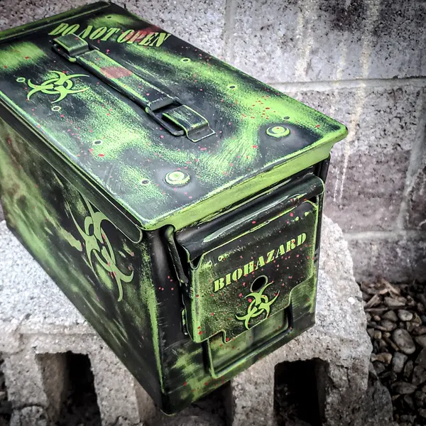 Steel ammo can coated in distressed green and black Cerakote with biohazard symbols and stencil lettering.